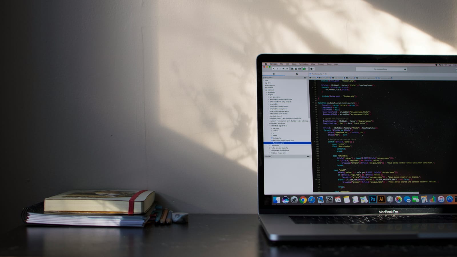 A laptop showing code beside a stack of notebooks on a desk, lit by soft window light