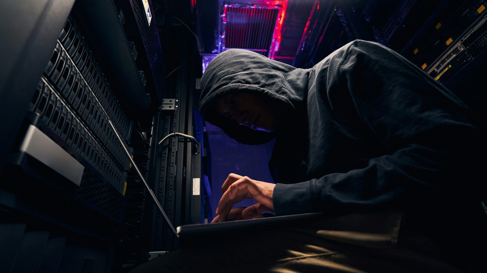A hooded figure at a laptop in a dark server room