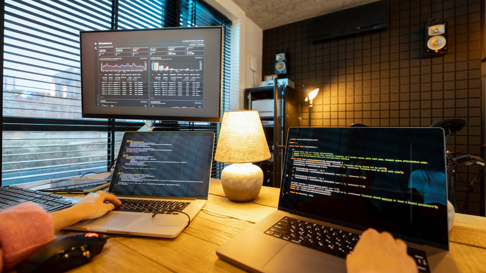 A home-office desk at night with two laptops showing code and a dashboard on a monitor